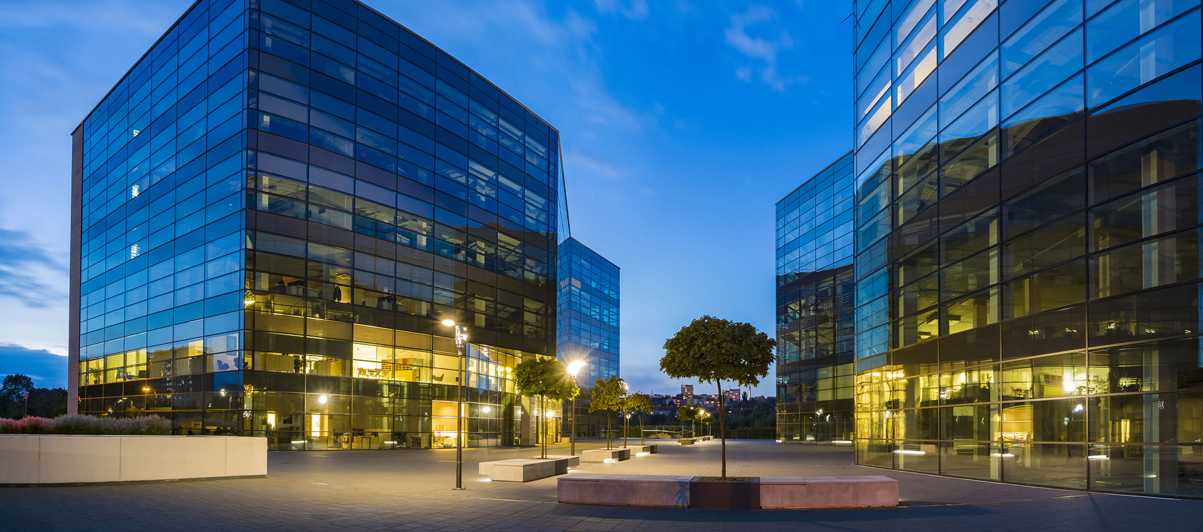The cityscape of four modern office buildings with glass facade at dusk, surrounded with trees, street lamps, and stone benches