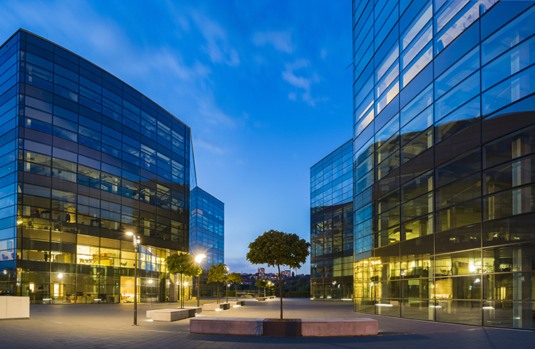 The cityscape of four modern office buildings with glass facade at dusk, surrounded with trees, street lamps, and stone benches