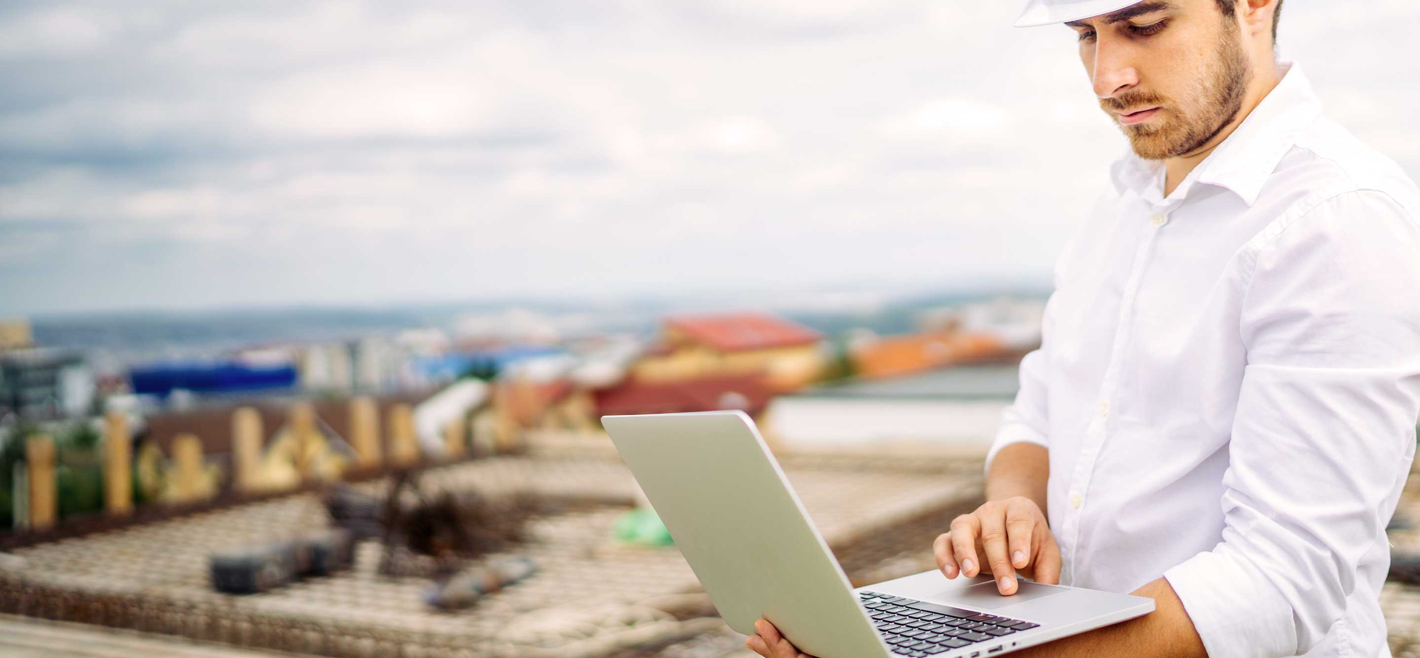 A construction manager wearing a white hard hat works on his laptop at a construction site