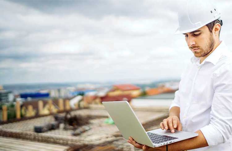 A construction manager wearing a white hard hat works on his laptop at a construction site