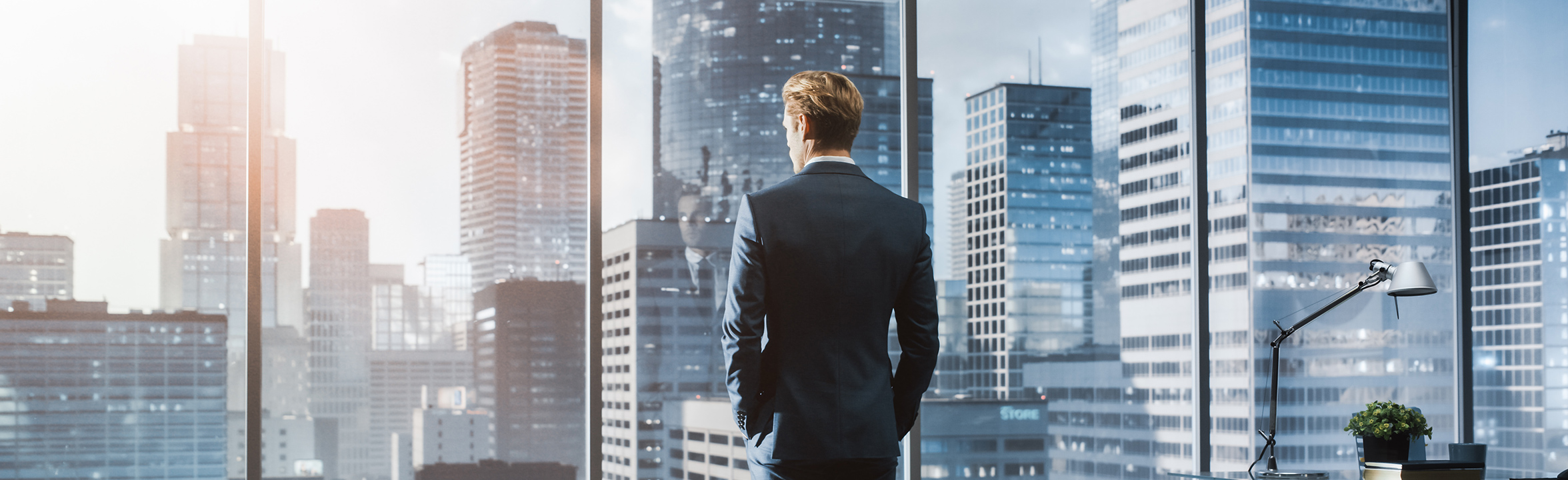 Young businessman in suit standing in modern office looking out of the window on city with skyscrapers