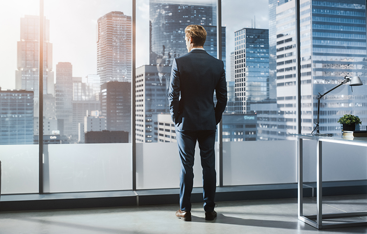 Young businessman in suit standing in modern office looking out of the window on city with skyscrapers