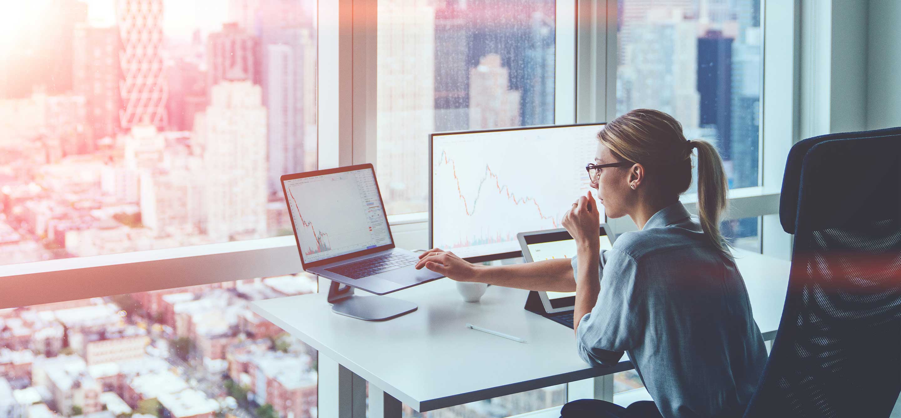 Eine Geschaeftsfrau arbeitet an ihrem Computer mit Finanzdaten in einem Buero mit Blick auf Wolkenkratzer