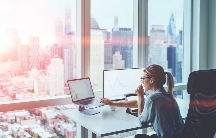 Eine Geschaeftsfrau arbeitet an ihrem Computer mit Finanzdaten in einem Buero mit Blick auf Wolkenkratzer